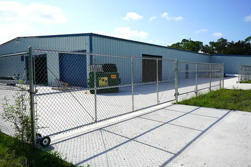 Galvanized chain link fence and rolling gate across the concrete drive of a DeBary commercial yard with a metal warehouse building behind it