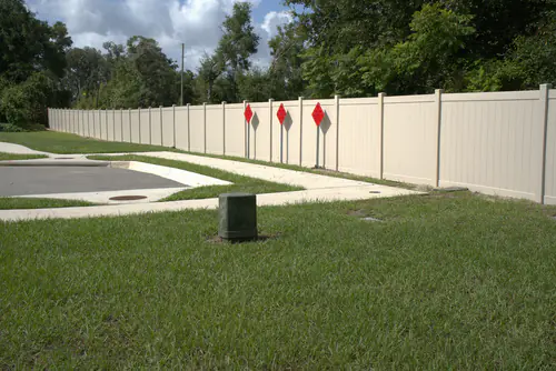 Long tan vinyl privacy fence running behind a sidewalk at the end of an Apopka road with red object markers and a utility pedestal in the grass shoulder