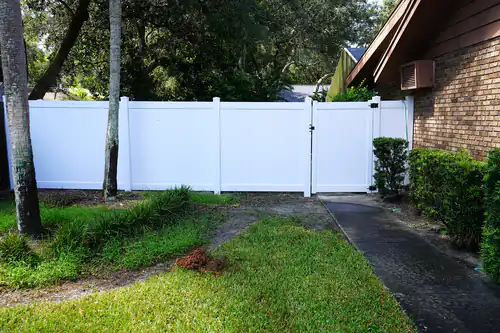 White vinyl privacy fence panels and a walk gate beside a Deltona driveway, with palm and oak trees close to the fence line